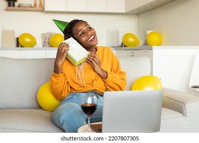 Overjoyed Black Woman Celebrating B-day Online Via Laptop, Holding Birthday Gift And Smiling, Sitting On Sofa, Having Video Call With Friends At Home. Virtual Holiday Celebration