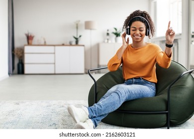 Overjoyed black woman wearing wireless headphones dancing sitting on beanbag chair, happy attractive young female listening to popular music, moving to favorite song, full body length, free copy space