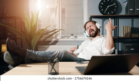 Overjoyed Businessman With Legs On Desk Listening To Music In Headphones Pretending To Play Guitar At Office