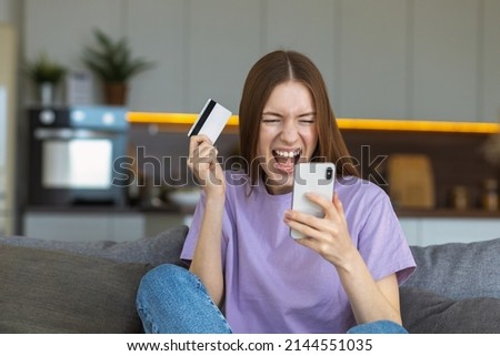 Overjoyed Caucasian woman with open mouth holding mobile phone and credit card sitting at home on the couch. Online shopping, technology concept