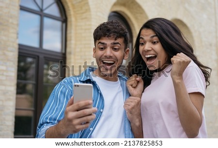 Overjoyed couple of friends using smartphone playing mobile game, celebration success. Emotional Indian man and African American woman shopping online with sales. Excited freelancers receive payment 