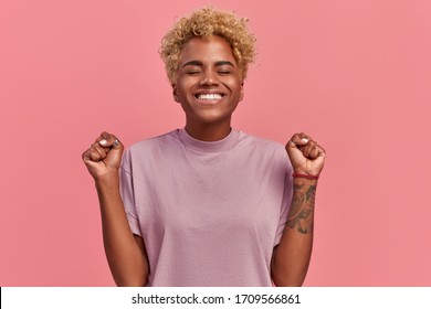 Overjoyed dark skinned woman makes victory dance, raises clenches fists and lifts up from joy, happy to receive awesome present or rejoicing triumph feeling winner, smiles broadly, on pink background.