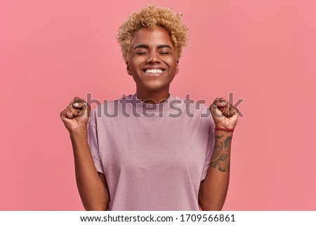 Overjoyed dark skinned woman makes victory dance, raises clenches fists and lifts up from joy, happy to receive awesome present or rejoicing triumph feeling winner, smiles broadly, on pink background.