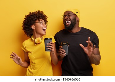 Overjoyed dark skinned woman with modern headphones laughs sincerely, dances together with Afro American boyfriend, drink hot coffee from disposable cups, stand together over yellow background