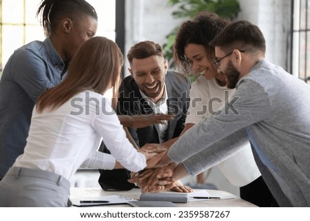 Overjoyed diverse businesspeople stack hands engaged in teambuilding in office together, excited multiracial colleagues coworkers involved in team motivational training at meeting, teamwork concept