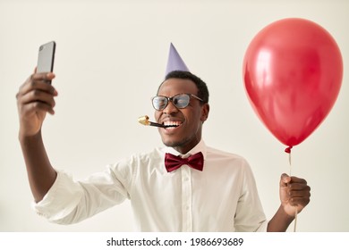 Overjoyed Ecstatic Young African Male In Elegant Clothing Blowing Party Horn, Holding Mobile Phone, Laughing Out Loud While Having Video Conference Call On His Birthday. Technology And Celebration