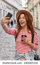 Overjoyed European Female Traveler In Hat, Makes Selfie Portrait Outdoor, Has Fun During Excursion In Ancient Town, Drinks Coffee From Disposable Cup, Wears Stylish Hat, Laughs Joyfully At Camera