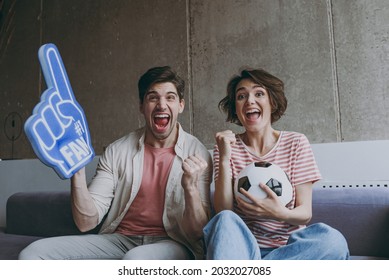 Overjoyed excited couple woman man friends football fans wearing casual clothes foam glove finger up support favorite team with soccer ball indoors at home watch tv People sport leisure family concept