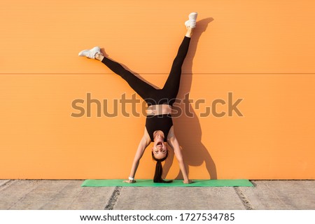 Overjoyed excited girl with perfect athletic body in tight sportswear doing yoga handstand pose against wall and laughing, shouting from happiness. Gymnastics for body balance outdoor workouts