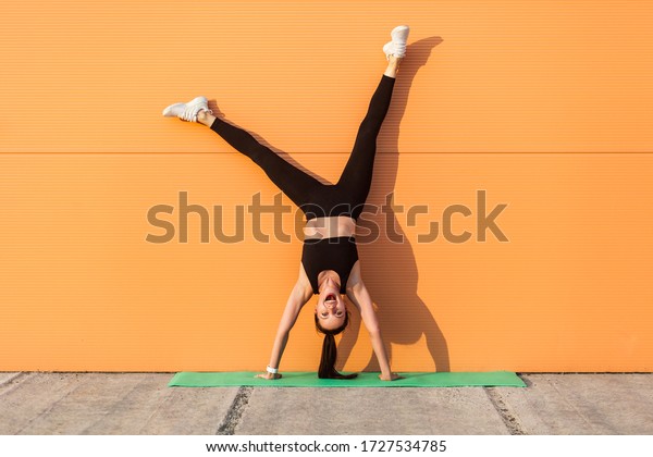 Overjoyed excited girl with perfect athletic\
body in tight sportswear doing yoga handstand pose against wall and\
laughing, shouting from happiness. Gymnastics for body balance\
outdoor workouts
