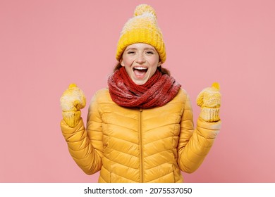 Overjoyed excited happy young woman 20s years old wears yellow jacket hat mittens doing winner gesture celebrate clenching fists say yes isolated on plain pastel light pink background studio portrait
