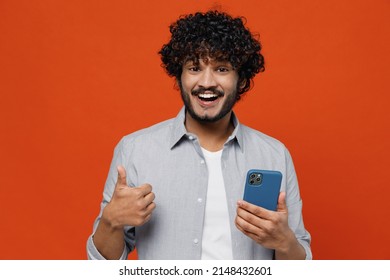 Overjoyed excited jubilant young bearded Indian man 20s years old wears blue shirt hold in hand use mobile cell phone showing thumb up like gesture isolated on plain orange background studio portrait