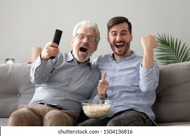 Overjoyed Excited Two Generations Men Family Old Senior Father And Young Adult Grown Son Hold Snack Remote Control Watching Tv Sport Game Match Scream Support Celebrate Team Victory At Home On Sofa