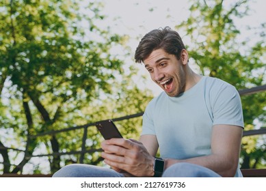 Overjoyed exultant young happy man 20s in blue t-shirt sit on bench using mobile cell phone chat online scream rest relax in sunshine spring green city park outdoor on nature Urban leisure concept. - Shutterstock ID 2127619736