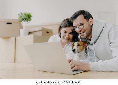 Overjoyed Family Couple Laugh As Watch Online Stores On Modern Laptop Computer With Their Dog, Have Break After Unpacking Carton Boxes, Home Renovation, Being In Good Mood. Newly Rented House.