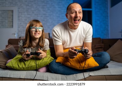 Overjoyed Father And Daughter Playing Video Games, Selective Focus. Happy Family Sitting On A Sofa And Playing Video Games With Blue Neon Light In Background. Smiling Family Sitting On The Couch 