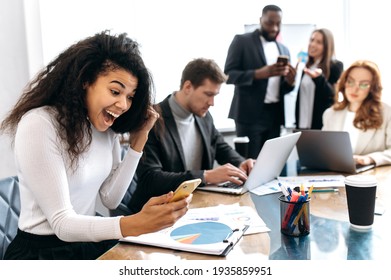 Overjoyed female employee is sitting in office, looking at smartphone screen, smiling. Excited african american business woman well done with project or startup, receive big profit, success concept