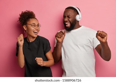 Overjoyed funny Afro American girl and her male friend sing along favourite track, dance with arms raised, move against pink background, wear black and white clothes, have happy moment in life - Shutterstock ID 1588906726