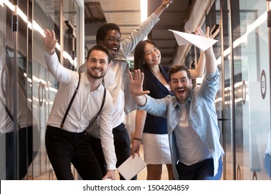Overjoyed funny multiracial millennial employees look at camera having fun in office hallway, happy excited multiethnic diverse work group, young motivated team posing for picture in corridor