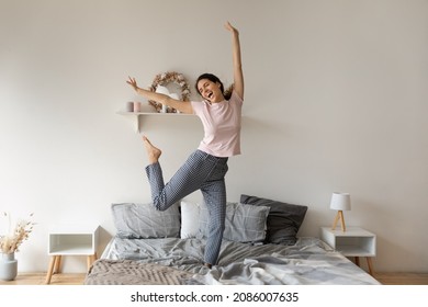 Overjoyed funny woman jumping on comfortable cozy bed at home, enjoying morning, starting new day, excited happy young female wearing pajama dancing to favorite music, having fun in modern bedroom