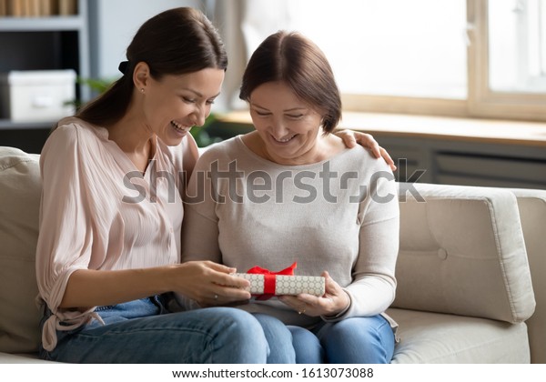 Overjoyed grownup daughter sit on couch with smiling\
senior mother greeting giving wrapped gift, caring adult girl child\
congratulate make birthday surprise to middle-aged mom, celebrating\
at home