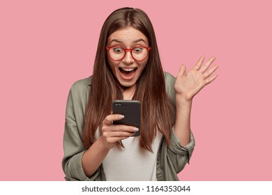 Overjoyed happy excited female glad to recieve text message informing about salary, rejoices good news, stares at mobile phone, gestures actively from happiness, stands indoor alone. Reaction concept