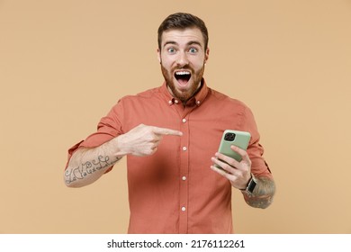 Overjoyed happy excited young brunet man 20s he wears orange shirt hold in hand use mobile cell phone points on it isolated on plain pastel light beige background studio portrait. Tattoo translate fun