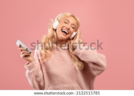 Overjoyed happy lady with headphones and smartphone listening to music and dancing, enjoying cool playlist and moving to favorite song over pink background
