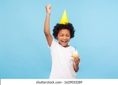 Overjoyed happy little boy with party cone holding cupcake and keeping eyes closed while making wish on his birthday, standing with raised hand shouting from happiness. studio shot blue background