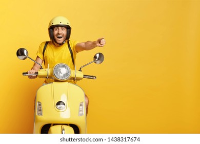 Overjoyed happy male drives motocycle, dressed in casual yellow t shirt and headgear, points happily into distance, notices something awesome, reaches destination on fast transport, enjoys speed