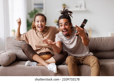 Overjoyed happy young african american couple man and woman raising arms, celebrating goal and screaming while watching football match together at home, selective focus. Leisure weekend activities