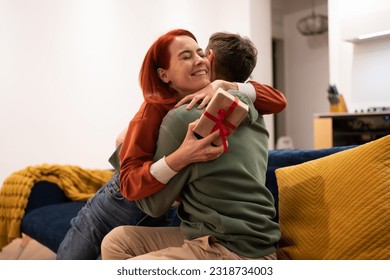 Overjoyed happy young woman holding gift box hugging husband feeling excited getting present from boyfriend. Family couple celebrating birthday and anniversary together at home, sitting on sofa - Shutterstock ID 2318734003
