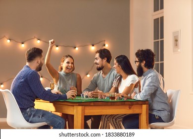 Overjoyed, Happy Young Woman Laughing And Raising Arm, Celebrating Her Victory In Game Of Poker, Sitting At Table With Friends At Casino Themed Party At Home Or Club