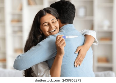 Overjoyed indian woman embracing man from behind, holding positive pregnancy test, both sharing moment of happiness and anticipation in bright room - Powered by Shutterstock