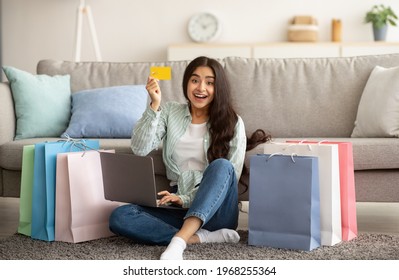 Overjoyed Indian Woman Feeling Happy Over Big Sale In Online Shop, Using Laptop, Showing Credit Card, Sitting On Floor With Gift Bags At Home. Excited Lady Purchasing Goods Over Internet Indoors