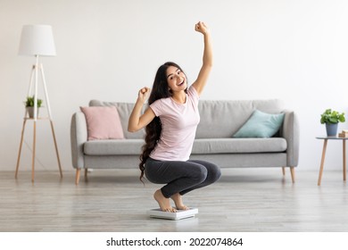 Overjoyed Indian woman sitting on scales, gesturing YES, excited over result of her weight loss diet at home. Millennial Asian lady achieving her slimming goal, copy space