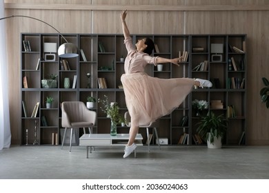 Overjoyed Indian woman wearing dress dancing, jumping in modern living room interior alone, having fun, listening to favorite good music, happy beautiful young female enjoying leisure time at home
