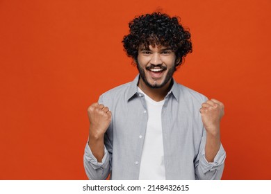 Overjoyed jubilant excited exultant young bearded Indian man 20s years old wears blue shirt doing winner gesture celebrate clenching fists say yes isolated on plain orange background studio portrait