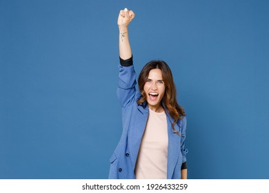 Overjoyed laughing screaming young brunette woman 20s in basic jacket standing rising hand clenching fist doing winner gesture looking camera isolated on bright blue colour background studio portrait
