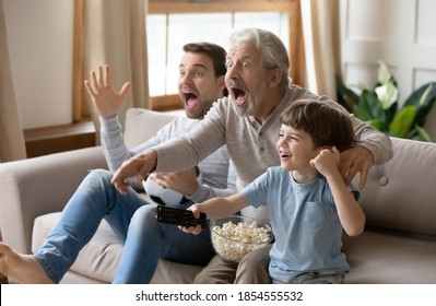 Overjoyed Little Boy With Father And Grandfather Celebrating Goal, Sitting On Couch With Popcorn And Soccer Ball, Excited Happy Three Generations Of Men Watching Tv, Supporting Favorite Football Team