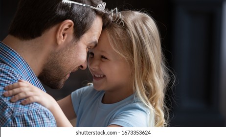 Overjoyed Little Preschooler Girl Child Touch Foreheads Look In Eyes Playing With Loving Young Dad Wearing Toy Crown, Happy Father And Small Daughter Have Fun Playing Together Funny Games At Home