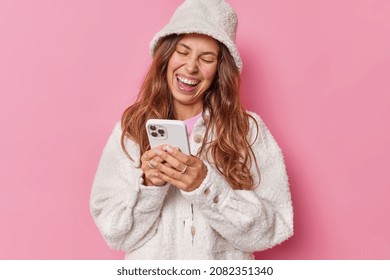 Overjoyed long haired woman uses mobile phone watches funny video wears fur hat and jacket views photos after party isolated over pink background. People positive emotions technology concept - Powered by Shutterstock