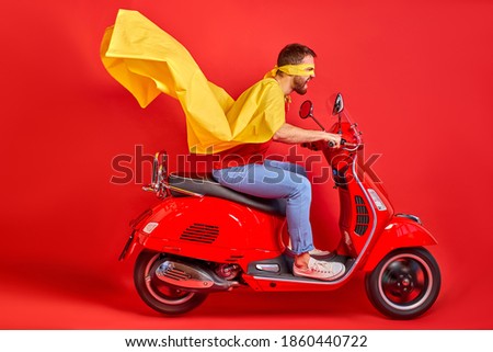 overjoyed mad crazy man driving moped fast speed wearing super hero costume isolated over red background, cloak sways in the wind