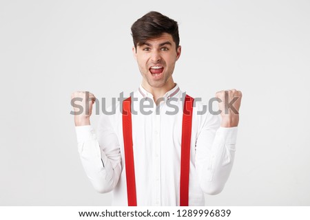 Overjoyed male clenches fists with happiness, opens mouth widely as shouts loudly, celebrates his success, poses against whitebackground. Happy stylish man with joyful expression