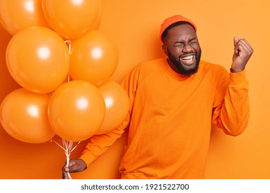 Overjoyed male student celebrates success makes yes gesture being on graduation party holds inflated balloons dressed in casual jumper isolated over orange background. People happiness concept