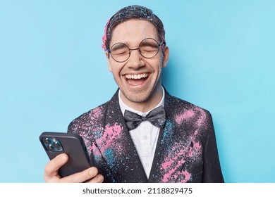 Overjoyed Male Student Enjoys University Leaving Party Wears Round Spectacles And Formal Suit Uses Cellphone For Chatting Laughs Joyfully Poses Against Blue Background. People Celebration Concept