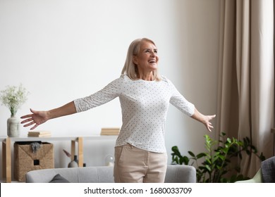 Overjoyed mature grandmother standing with outstretched arms near comfortable couch, breathing fresh air, enjoying freedom, happy life moment. Smiling older woman feeling thankful for good day.