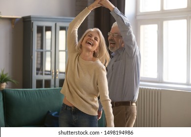 Overjoyed mature grey-haired Caucasian husband and wife have fun enjoy time together at home, happy elderly couple spouses dancing in living room, senior man lead sway smiling middle-aged woman