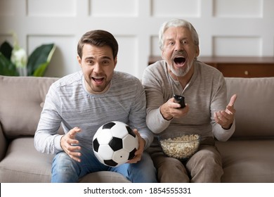 Overjoyed Mature Man Father With Adult Son Watching Tv, Match Together, Excited Shouting Older Grandfather With Grandson Sitting On Couch With Popcorn And Soccer, Looking At Camera, Supporting Team