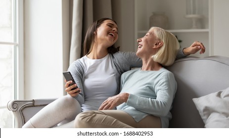 Overjoyed Mature Mother And Millennial Grown-up Daughter Relax On Sofa In Living Room Laugh Watch Funny Video On Smartphone, Happy Senior Mom And Adult Girl Child Have Fun Using Cellphone Together